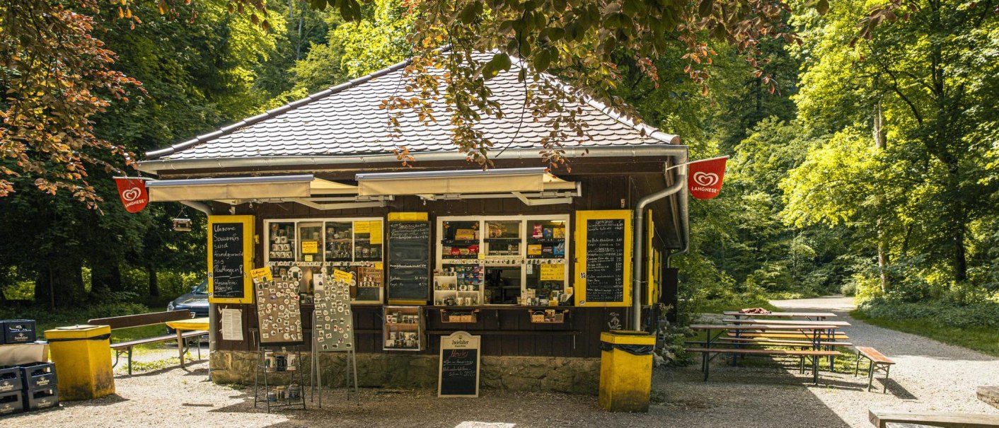 Ein Kiosk im Wald mit Verkaufstafeln und Sitzbänken. Langnese-Flaggen hängen an den Seiten. Der Weg führt durch die grüne Umgebung., © Stuttgart-Marketing GmbH, Sarah Schmid