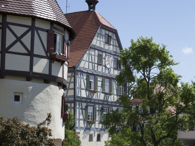 Fachwerkh&auml;user in Merklingen, umgeben von gr&uuml;nen B&auml;umen und blauem Himmel. Traditionelle Architektur mit roten D&auml;chern und Holzverzierungen., &copy; Stadtverwaltung Weil der Stadt
