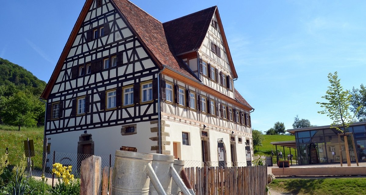 Fachwerkhaus mit spitzem Dach, umgeben von Natur. Im Vordergrund ein Holzzaun mit Gießkannen, im Hintergrund blauer Himmel und ein modernes Gebäude., © Bildarchiv Freilichtmuseum Beuren Fachwerkhaus mit spitzem Dach, umgeben von Natur. Im Vordergrund ein Holzzaun mit Gießkannen, im Hintergrund blauer Himmel und ein modernes Gebäude., © Bildarchiv Freilichtmuseum Beuren