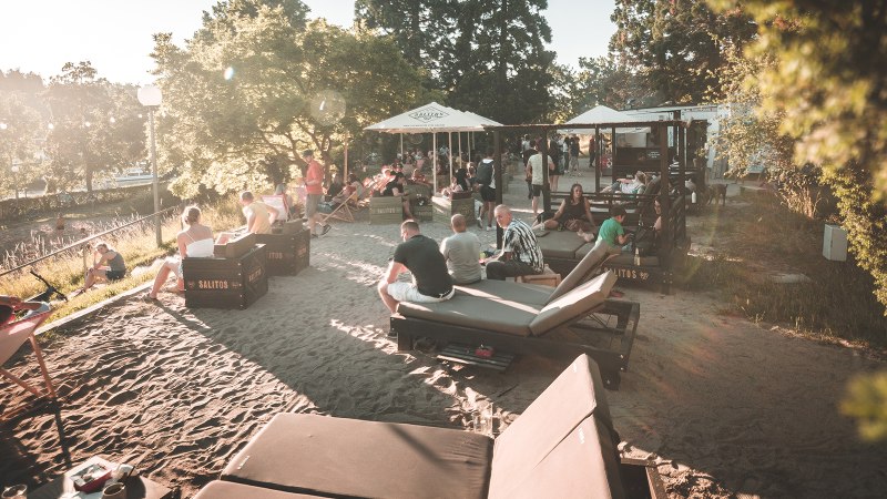Menschen entspannen an einem sonnigen Stadtstrand in Stuttgart. Liegestühle und Sonnenschirme bieten eine gemütliche Atmosphäre im Sand., © Classic Rock Café Gastronomie- Betriebs GmbH Menschen entspannen an einem sonnigen Stadtstrand in Stuttgart. Liegestühle und Sonnenschirme bieten eine gemütliche Atmosphäre im Sand., © Classic Rock Café Gastronomie- Betriebs GmbH