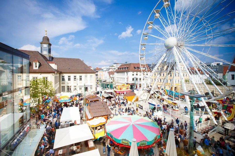 Ein belebter Platz in G&ouml;ppingen mit einem gro&szlig;en Riesenrad, bunten St&auml;nden und vielen Menschen bei sonnigem Wetter.
