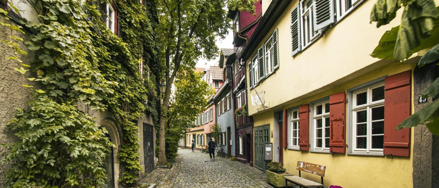 Eine charmante, gepflasterte Gasse in Esslingen, gesäumt von bunten Fachwerkhäusern und üppigem Grün. Zwei Personen spazieren die Straße entlang., © SMG, Sarah Schmid Eine charmante, gepflasterte Gasse in Esslingen, gesäumt von bunten Fachwerkhäusern und üppigem Grün. Zwei Personen spazieren die Straße entlang., © SMG, Sarah Schmid