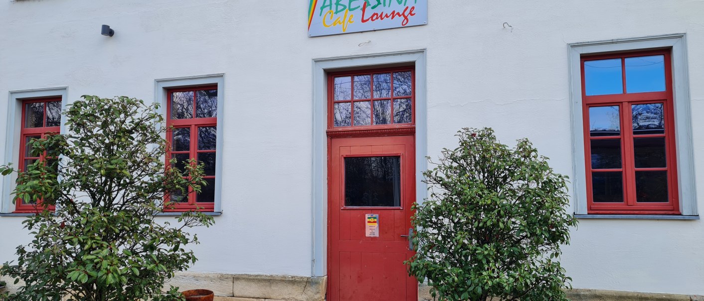 Der Eingangsbereich der Abessina Caf&eacute; Lounge mit roter T&uuml;r und Fenstern, flankiert von zwei Pflanzen in K&uuml;beln., &copy; cAbessina