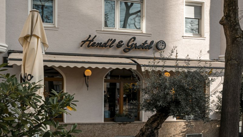 Das Bild zeigt die Fassade des Restaurants Hendl & Gretel mit einem Sonnenschirm und Pflanzen im Vordergrund., © SMG, Sarah Schmid Das Bild zeigt die Fassade des Restaurants Hendl & Gretel mit einem Sonnenschirm und Pflanzen im Vordergrund., © SMG, Sarah Schmid