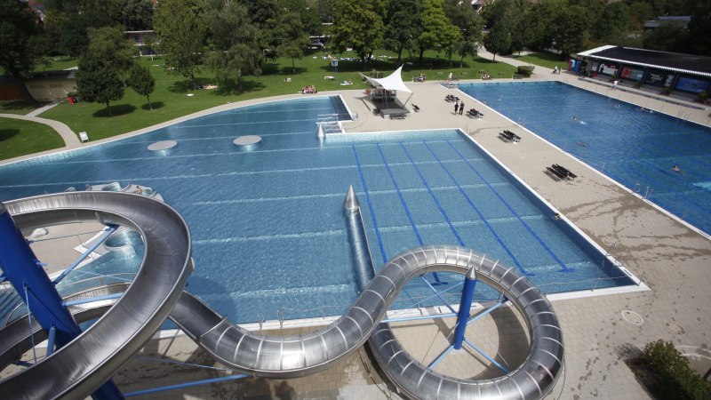 Freibad Rosental mit großer Wasserrutsche, mehreren Schwimmbecken und umliegenden Grünflächen. Menschen entspannen auf der Wiese., © Stuttgarter Bäder