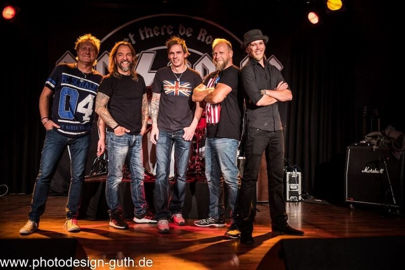 F&uuml;nf Bandmitglieder posieren auf einer B&uuml;hne vor einem AC/DC-Logo. Sie tragen l&auml;ssige Kleidung und l&auml;cheln in die Kamera., &copy; EMT Event-Media-Tec GmbH