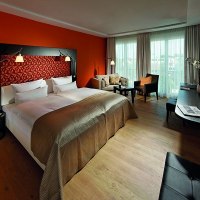 Modernes Hotelzimmer mit großem Bett, oranger Wand, Sofa, Schreibtisch und Fernseher. Helle Vorhänge und Holzboden schaffen eine gemütliche Atmosphäre., © Parkhotel Stuttgart Messe-Airport