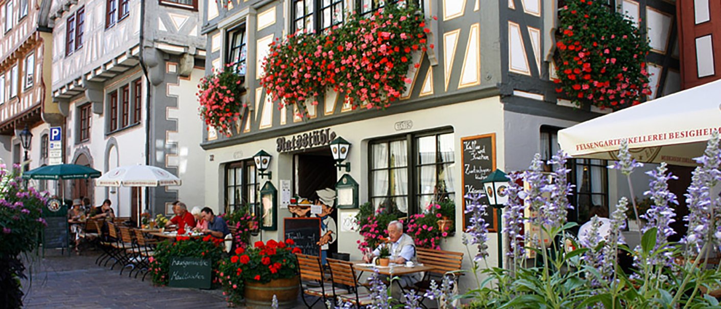 Fachwerkhaus in Besigheim mit Restaurant 'Ratsstüble'. Menschen sitzen draußen unter Sonnenschirmen, umgeben von blühenden Blumen., © Ratsstüble Besigheim