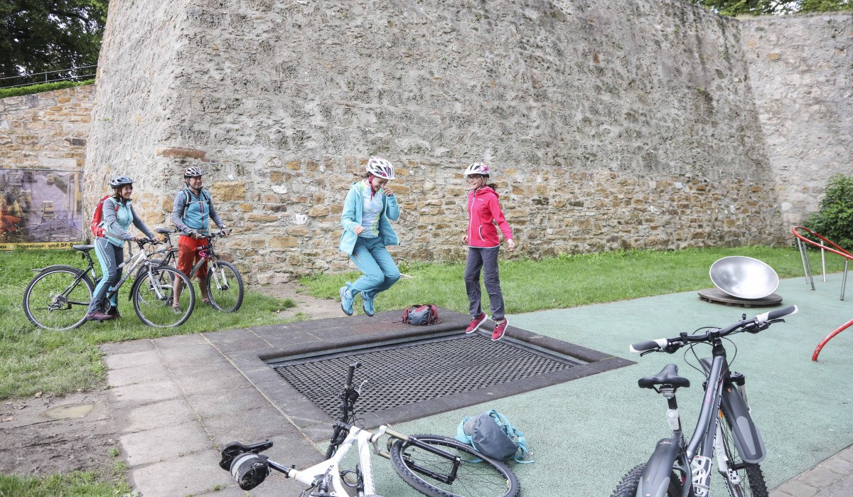 Zwei Personen springen auf einem Trampolin neben Fahrrädern vor einer alten Steinmauer. Weitere Personen stehen mit Fahrrädern daneben., © Schwäbische Alb Tourismusverband e.V.