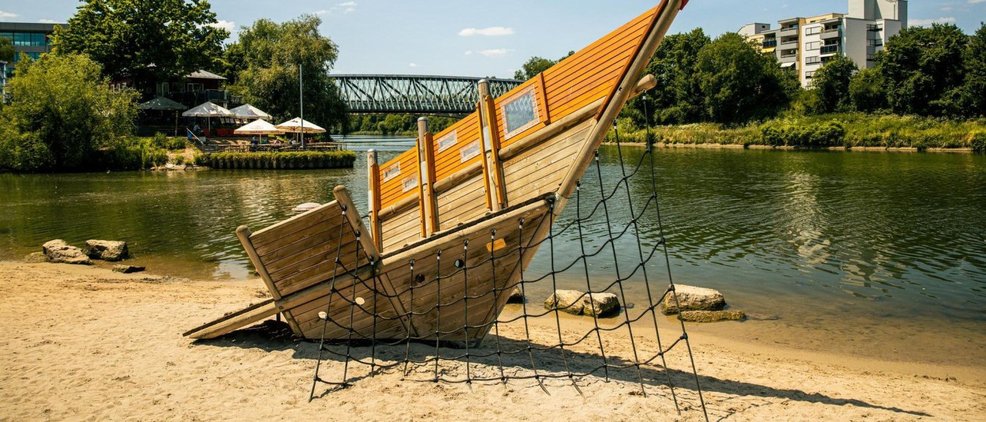 Ein hölzernes Spielschiff am Sandstrand des Neckars in Remseck. Im Hintergrund sind eine Brücke und Gebäude zu sehen., © Stuttgart-Marketing GmbH, Sarah Schmid Ein hölzernes Spielschiff am Sandstrand des Neckars in Remseck. Im Hintergrund sind eine Brücke und Gebäude zu sehen., © Stuttgart-Marketing GmbH, Sarah Schmid