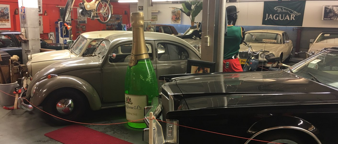 Innenansicht eines Museums mit Oldtimern, einem großen Champagnerflaschen-Modell und einem Jaguar-Logo an der Wand.