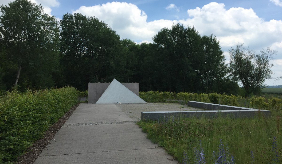 Ein Weg führt zu einem pyramidenförmigen Denkmal in einer Gedenkstätte, umgeben von Bäumen und Wiesen unter einem bewölkten Himmel., © www.pro-cycl.de