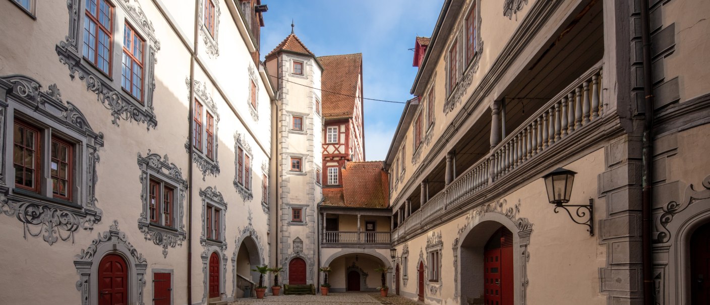 gepflasterter Schlossinnenhof, umgeben von hell verputzten Gebäuden mit grauer Illusionsmalerei umd Fenster und Türen, auf der rechten Bildseite eine offene Galerie im ersten Obergeschoss des Gebäudes, © stadt Gaildorf