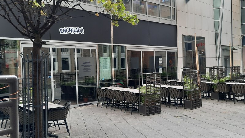 Außenbereich eines Restaurants namens Enchilada mit leeren Tischen und Stühlen. Ein Baum und Pflanzen trennen die Sitzbereiche., © SMG Außenbereich eines Restaurants namens Enchilada mit leeren Tischen und Stühlen. Ein Baum und Pflanzen trennen die Sitzbereiche., © SMG