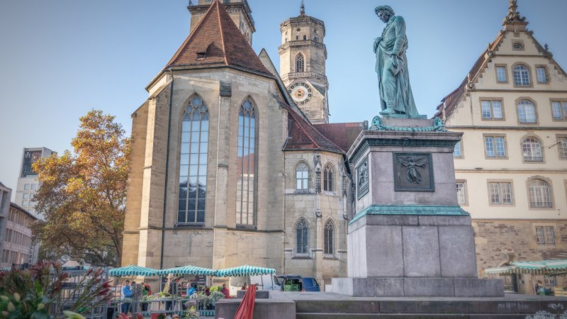 Der Schillerplatz in Stuttgart zeigt eine Statue, historische Gebäude und Marktstände. Im Hintergrund ist eine Kirche mit Turm zu sehen., © SMG Martina Denker
