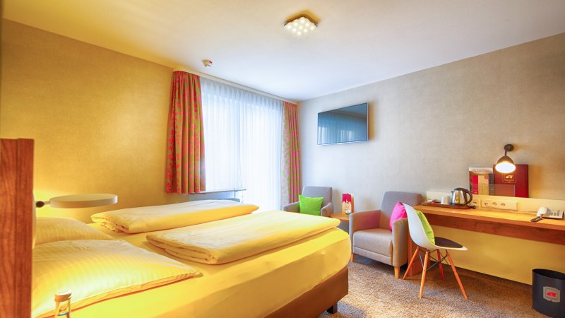 Ein gem&uuml;tliches Hotelzimmer mit Doppelbett, Schreibtisch, Fernseher und zwei Sesseln. Helle Vorh&auml;nge und bunte Kissen sorgen f&uuml;r eine einladende Atmosph&auml;re., &copy; TOMAS