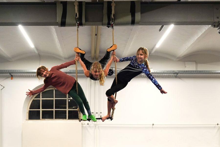 Drei Kinder hängen an Seilen in einer Halle und üben Luftakrobatik. Sie tragen sportliche Kleidung und wirken konzentriert und fröhlich., © Kunstschule Labyrinth Drei Kinder hängen an Seilen in einer Halle und üben Luftakrobatik. Sie tragen sportliche Kleidung und wirken konzentriert und fröhlich., © Kunstschule Labyrinth