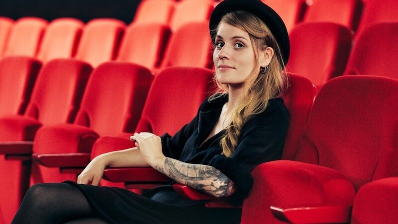 Eine Person mit Hut und tätowiertem Arm sitzt entspannt in einem leeren Kinosaal auf einem roten Sitz., © Mauerwerk GmbH Eine Person mit Hut und tätowiertem Arm sitzt entspannt in einem leeren Kinosaal auf einem roten Sitz., © Mauerwerk GmbH