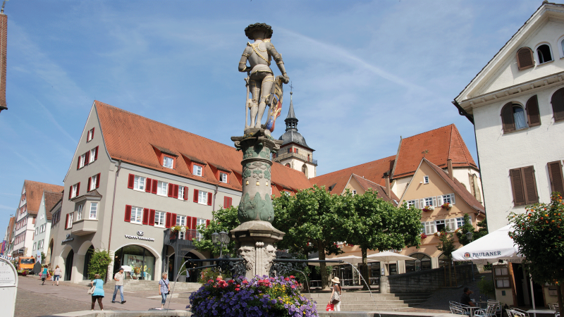 Marktbrunnen mit Statue auf einem belebten Platz, umgeben von historischen Gebäuden mit roten Dächern und Bäumen., © Stuttgart-Marketing GmbH Marktbrunnen mit Statue auf einem belebten Platz, umgeben von historischen Gebäuden mit roten Dächern und Bäumen., © Stuttgart-Marketing GmbH