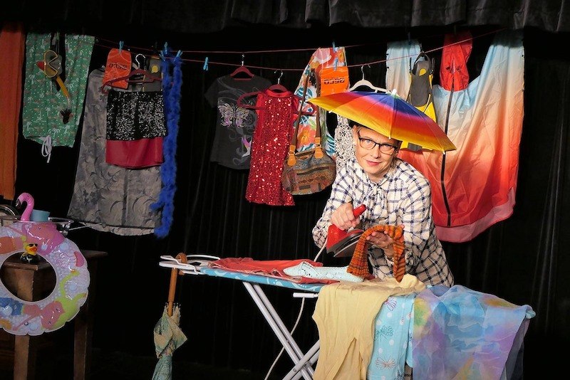 Eine Person b&uuml;gelt auf einer B&uuml;hne, umgeben von bunten Kleidungsst&uuml;cken und einem Regenschirm. Die Szene wirkt humorvoll und kreativ., &copy; Galli Theater Backnang