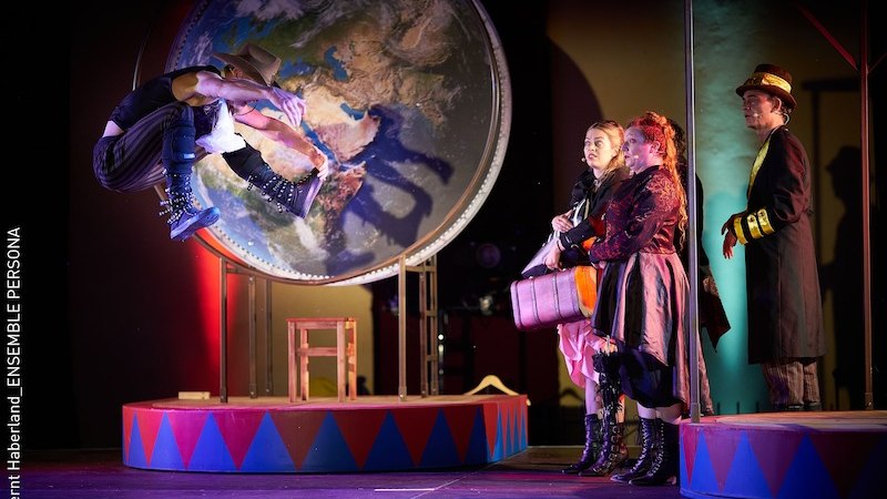 Theaterszene mit einem Akteur, der vor einer großen Weltkarte springt. Drei Personen in Kostümen beobachten ihn. Bühnenbild im Zirkusstil., © Stadt Kornwestheim Kulturmanagement Theaterszene mit einem Akteur, der vor einer großen Weltkarte springt. Drei Personen in Kostümen beobachten ihn. Bühnenbild im Zirkusstil., © Stadt Kornwestheim Kulturmanagement