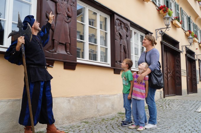Ein Stadtführer in historischer Kleidung zeigt einer Frau und zwei Kindern ein Relief an einer Hauswand in einer gepflasterten Straße., © Stadt Nürtingen Ein Stadtführer in historischer Kleidung zeigt einer Frau und zwei Kindern ein Relief an einer Hauswand in einer gepflasterten Straße., © Stadt Nürtingen