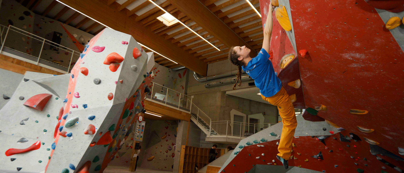 Eine Person klettert an einer roten Boulderwand in einer Indoor-Kletterhalle. Die Halle hat eine Holzdecke und verschiedene Kletterrouten., © Roccadion, Böblingen Eine Person klettert an einer roten Boulderwand in einer Indoor-Kletterhalle. Die Halle hat eine Holzdecke und verschiedene Kletterrouten., © Roccadion, Böblingen