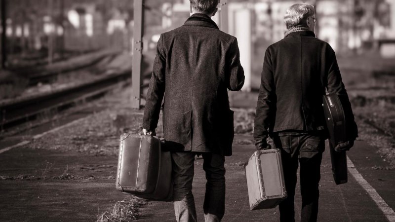 Zwei Männer in Mänteln tragen Koffer und gehen auf einem verlassenen Bahnsteig entlang. Die Szene ist in Schwarz-Weiß gehalten., © FMS Photography Zwei Männer in Mänteln tragen Koffer und gehen auf einem verlassenen Bahnsteig entlang. Die Szene ist in Schwarz-Weiß gehalten., © FMS Photography
