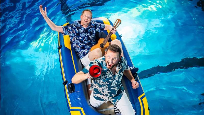 Zwei Männer in Hawaiihemden sitzen in einem Schlauchboot im Pool. Einer hält eine Gitarre, der andere ein Megafon. Sie wirken fröhlich und ausgelassen., © Dieselstrasse Kulturzentrum e.V.