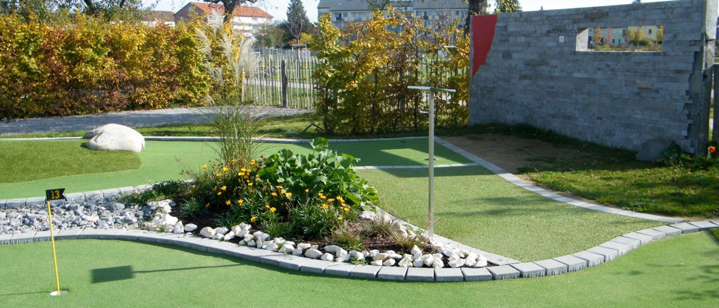Ein Minigolfplatz mit Loch 13, umgeben von Pflanzen und Steinen. Im Hintergrund sind Bäume und ein Zaun zu sehen., © Anja Beyerle & Ralf Bähr GbR