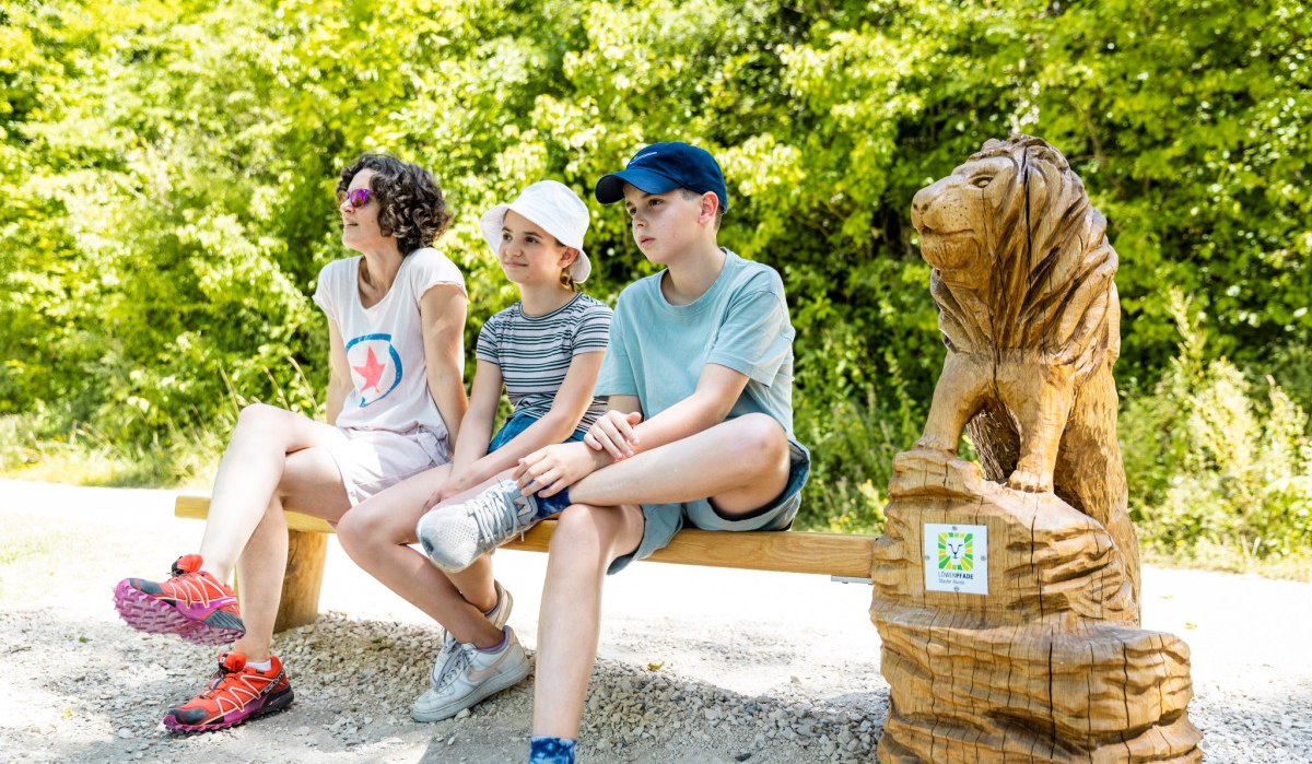 Drei Personen sitzen auf einer Holzbank neben einer geschnitzten Löwenskulptur im Grünen., © TMBW