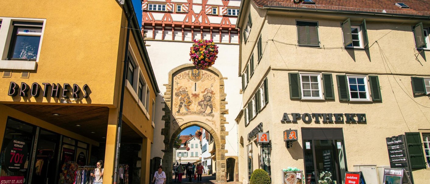 Das Stadttor von Bietigheim-Bissingen mit Fachwerk und Wandmalerei, umgeben von Geschäften wie einer Apotheke und einem Bekleidungsgeschäft., © Stuttgart-Marketing GmbH, Sarah Schmid Das Stadttor von Bietigheim-Bissingen mit Fachwerk und Wandmalerei, umgeben von Geschäften wie einer Apotheke und einem Bekleidungsgeschäft., © Stuttgart-Marketing GmbH, Sarah Schmid