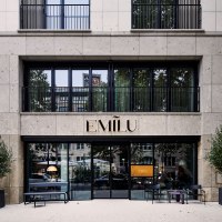 Eingang des EmiLu Hotels mit gro&szlig;en Glasfenstern, Pflanzen in schwarzen T&ouml;pfen und dem Hotelnamen &uuml;ber der T&uuml;r., &copy; EmiLu Hotel GmbH