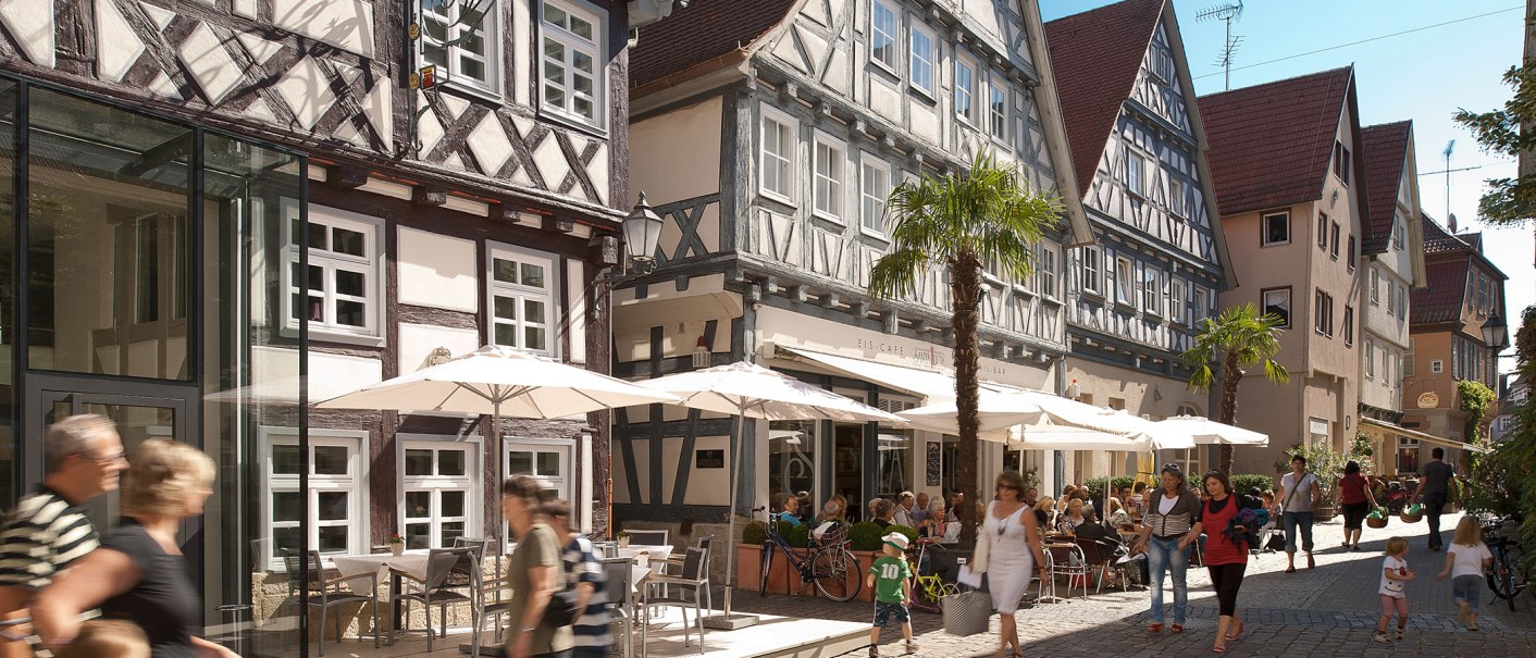 Belebte Straße mit Fachwerkhäusern, Cafés und Passanten. Menschen sitzen unter Sonnenschirmen, während andere vorbeigehen. Palmen schmücken die Szene., © Oswald Belebte Straße mit Fachwerkhäusern, Cafés und Passanten. Menschen sitzen unter Sonnenschirmen, während andere vorbeigehen. Palmen schmücken die Szene., © Oswald