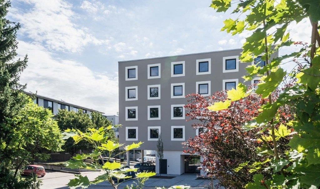 Modernes Hotelgeb&auml;ude mit quadratischen Fenstern, umgeben von gr&uuml;nen und roten Pflanzen, unter blauem Himmel., &copy; a2 HOTELS am Quadrium