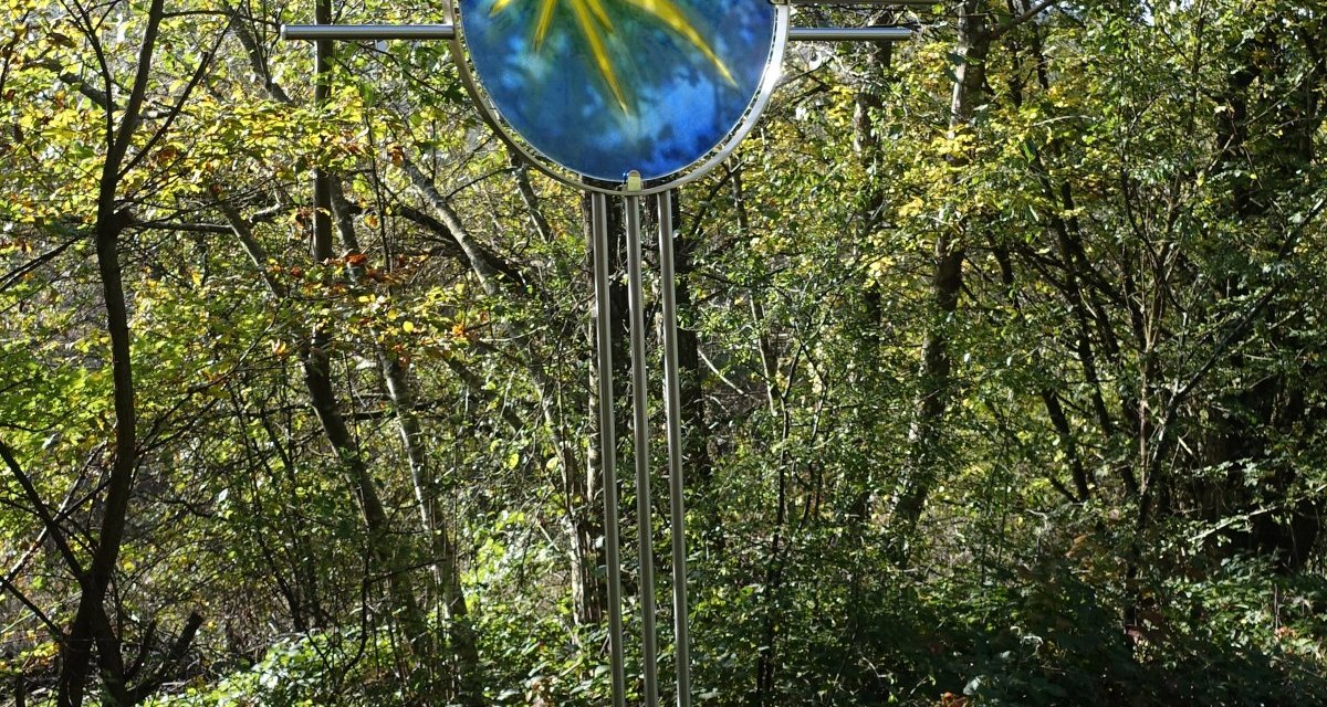 Ein Kreuz mit einem blauen und gelben Glasornament steht in einem Wald, umgeben von Bäumen und Pflanzen., © Foto: Cornelia Steinbach Ein Kreuz mit einem blauen und gelben Glasornament steht in einem Wald, umgeben von Bäumen und Pflanzen., © Foto: Cornelia Steinbach