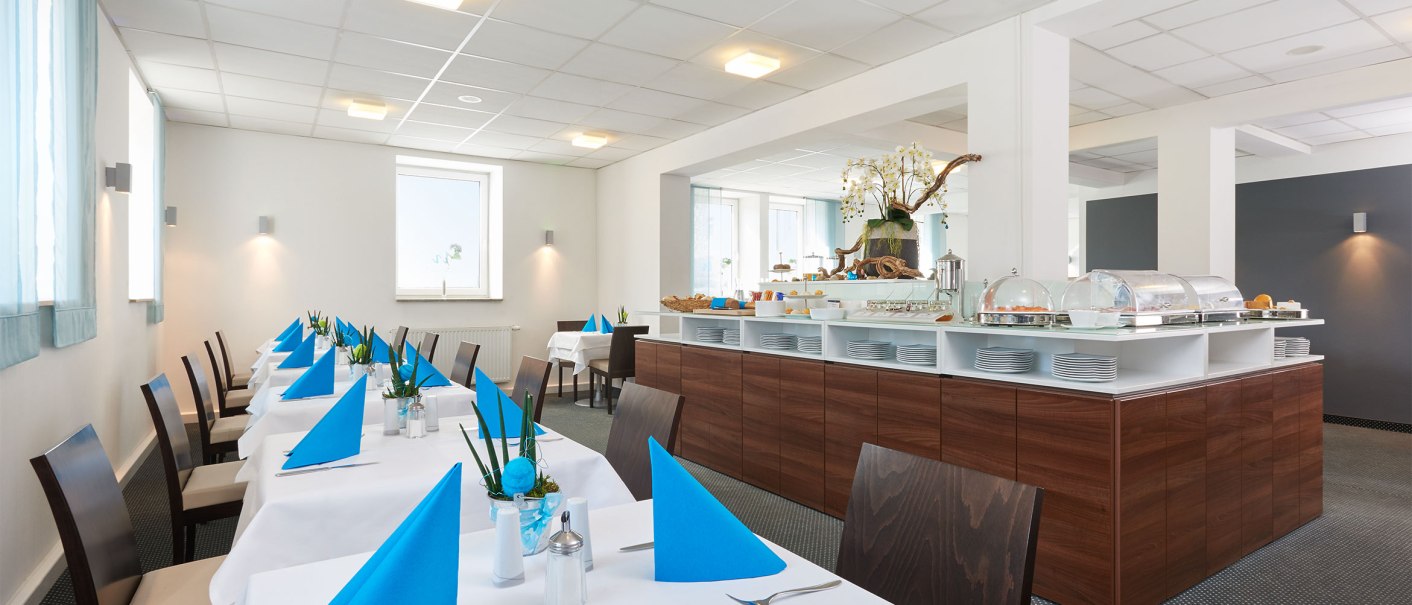 Ein heller Frühstücksraum mit weiß gedeckten Tischen, blauen Servietten und einem Buffet im Hintergrund., © TOMAS