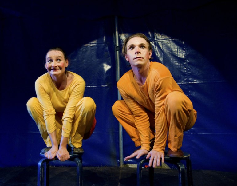 Zwei Personen in gelben und orangefarbenen Kostümen hocken auf Hockern vor einem blauen Hintergrund, wirken konzentriert und fröhlich., © Stadt Leinfelden-Echterdingen, Kulturamt