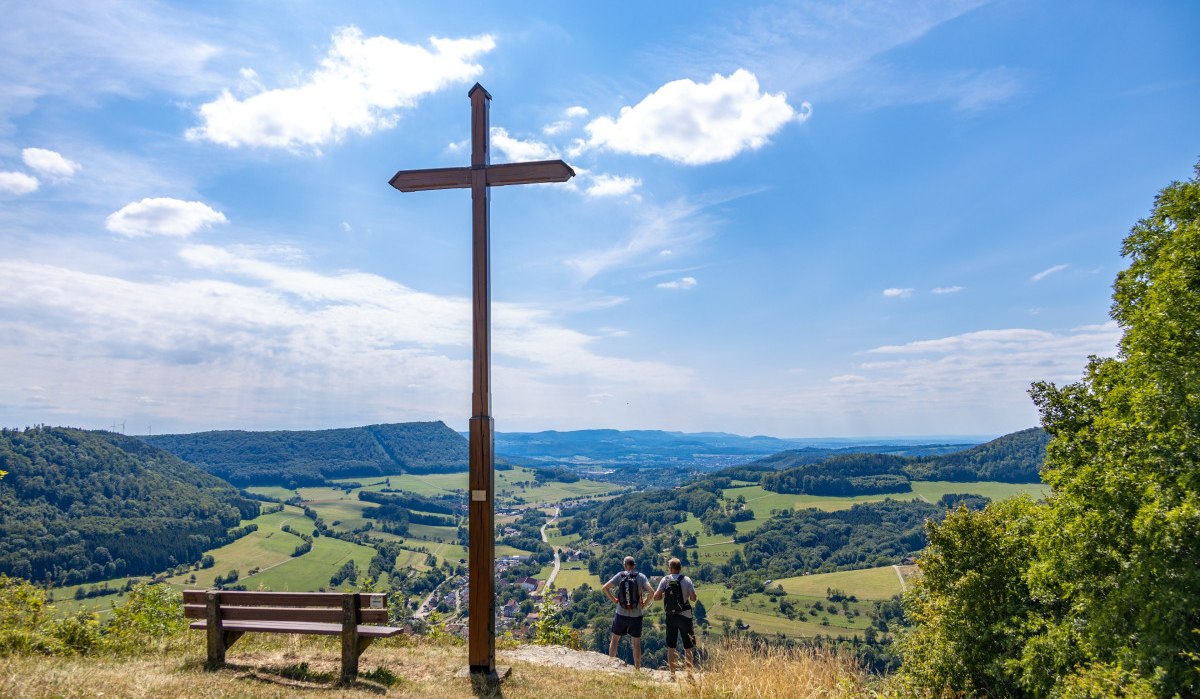 Gipfelkreuz auf einem Hügel mit Bank, zwei Personen blicken auf die weite Landschaft. Der Himmel ist blau mit einigen Wolken., © Foto Thomas Zehnder
