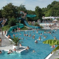 Ein belebtes Freibad mit zahlreichen Menschen im Wasser und auf den Rutschen. Umgeben von Bäumen und Pflanzen, herrscht eine sommerliche Atmosphäre., © Freibad Oberstenfeld
