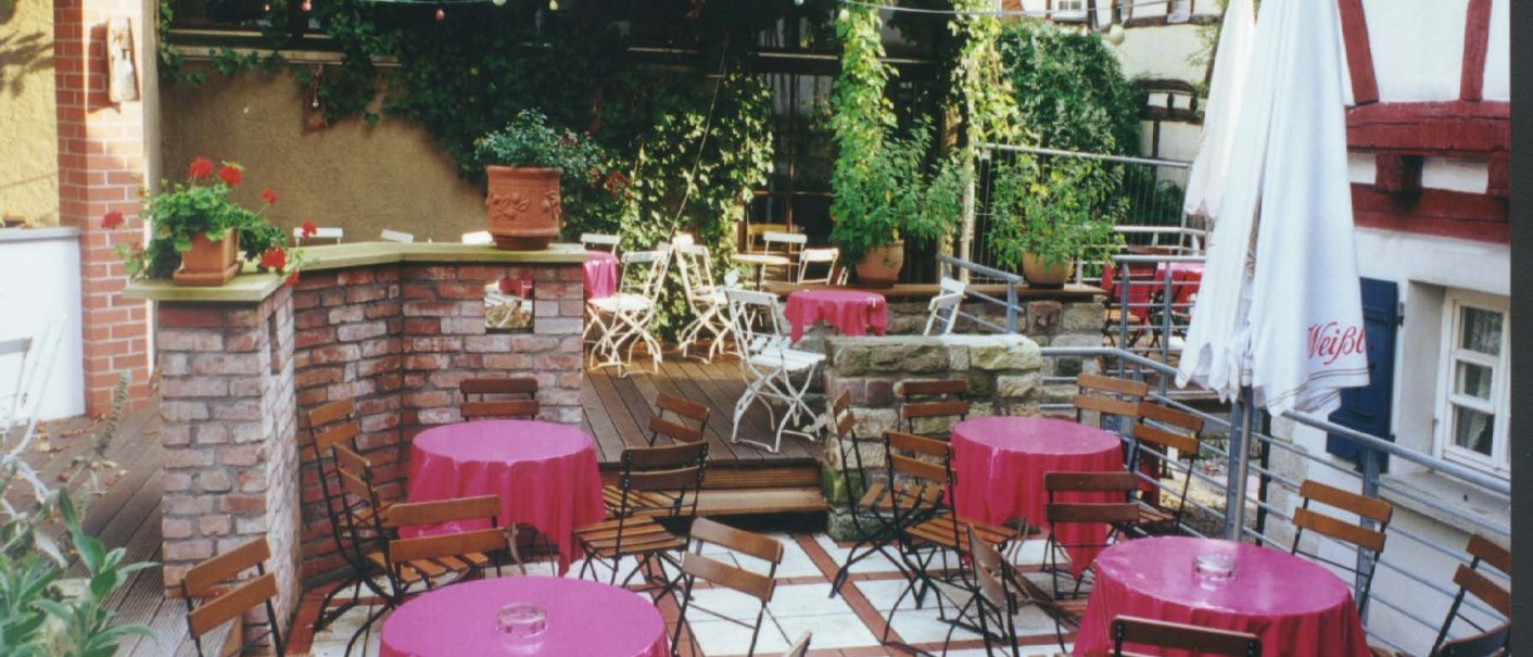 Ein gemütlicher Café-Innenhof mit Holztischen und -stühlen, rosa Tischdecken, umgeben von Pflanzen und Fachwerkhäusern., © 's Café