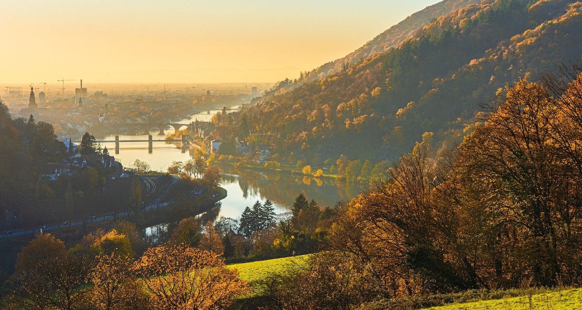 Herbstliche Landschaft am Neckar mit Br&uuml;cke und Stadt im Hintergrund, umgeben von H&uuml;geln und B&auml;umen im goldenen Licht der Abendd&auml;mmerung.