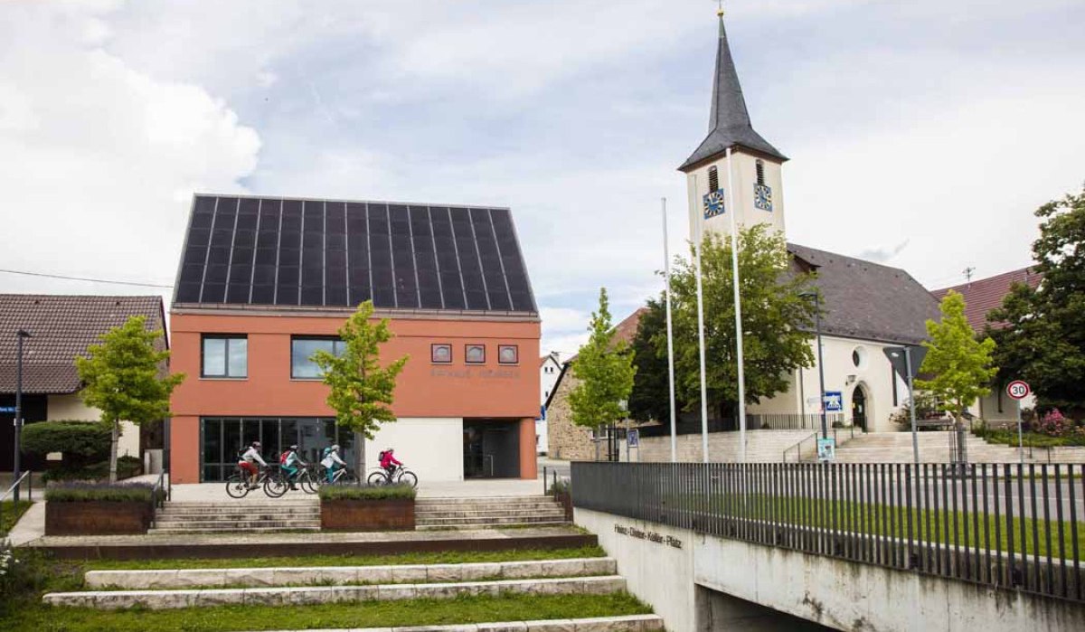 Ein modernes Gebäude mit Solarpanelen und eine Kirche in Jesingen. Vor dem Gebäude fahren Kinder auf Fahrrädern. Im Vordergrund sind Treppenstufen., © Schwäbische Alb Tourismusverband e.V. Ein modernes Gebäude mit Solarpanelen und eine Kirche in Jesingen. Vor dem Gebäude fahren Kinder auf Fahrrädern. Im Vordergrund sind Treppenstufen., © Schwäbische Alb Tourismusverband e.V.