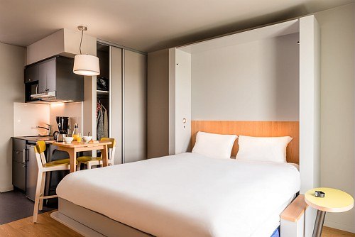 Modernes Studio mit großem Bett, kleiner Küche und Esstisch. Helle, minimalistische Einrichtung mit Holz- und Grautönen., © adagio access Stuttgart Airport Messe