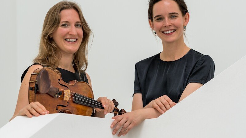 Zwei Frauen l&auml;cheln hinter einer wei&szlig;en Wand, eine h&auml;lt eine Geige. Sie tragen elegante, dunkle Kleidung., &copy; Mauerwerk GmbH