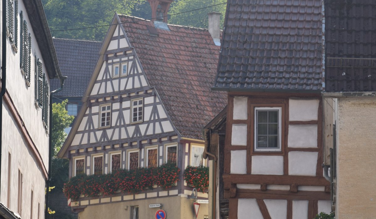 Fachwerkhäuser in einer engen, kurvigen Straße mit Blumen an den Fenstern und einem Hügel im Hintergrund., © Land der 1000 Hügel - Kraichgau-Stromberg Fachwerkhäuser in einer engen, kurvigen Straße mit Blumen an den Fenstern und einem Hügel im Hintergrund., © Land der 1000 Hügel - Kraichgau-Stromberg