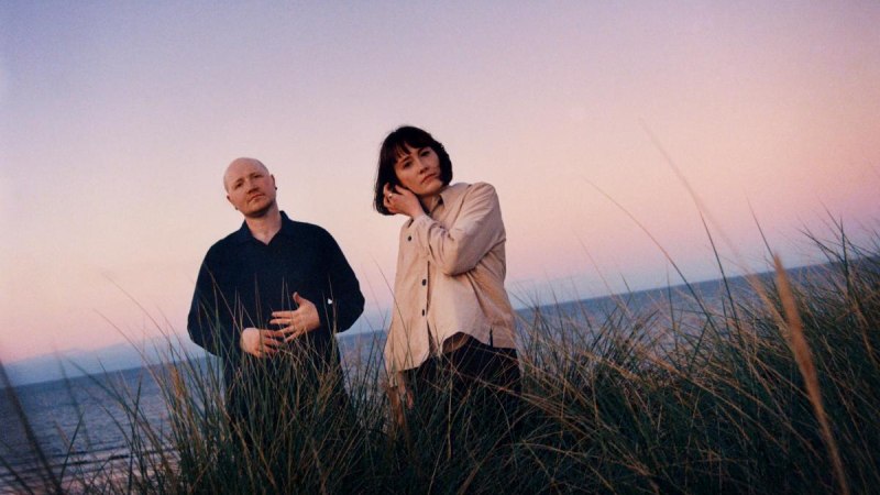 Zwei Personen stehen in hohen Gr&auml;sern an einem Strand. Der Himmel ist rosa gef&auml;rbt, was auf einen Sonnenuntergang hindeutet., &copy; Kulturzentrum Dieselstrasse