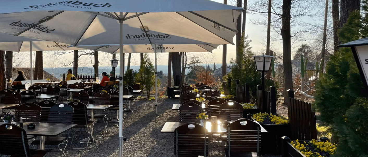 Ein ruhiger Biergarten mit Holztischen und Stühlen unter Sonnenschirmen, umgeben von Bäumen. Einige Gäste sitzen und genießen die Aussicht., © Schönbuch Waldbiergarten am Wasserturm Ein ruhiger Biergarten mit Holztischen und Stühlen unter Sonnenschirmen, umgeben von Bäumen. Einige Gäste sitzen und genießen die Aussicht., © Schönbuch Waldbiergarten am Wasserturm