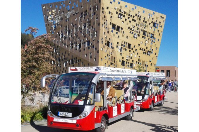 Zwei rote Elektrofahrzeuge fahren vor einem modernen, goldfarbenen Gebäude mit unregelmäßigen Fenstern. Menschen sitzen in den Fahrzeugen und genießen die Fahrt., © Touristik und Marketing GmbH