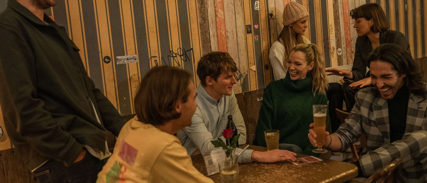 Eine Gruppe von Menschen sitzt in einer gemütlichen Bar und lacht. Sie genießen Getränke und unterhalten sich lebhaft. Der Raum hat eine warme Atmosphäre., © SMG, Martina Denker